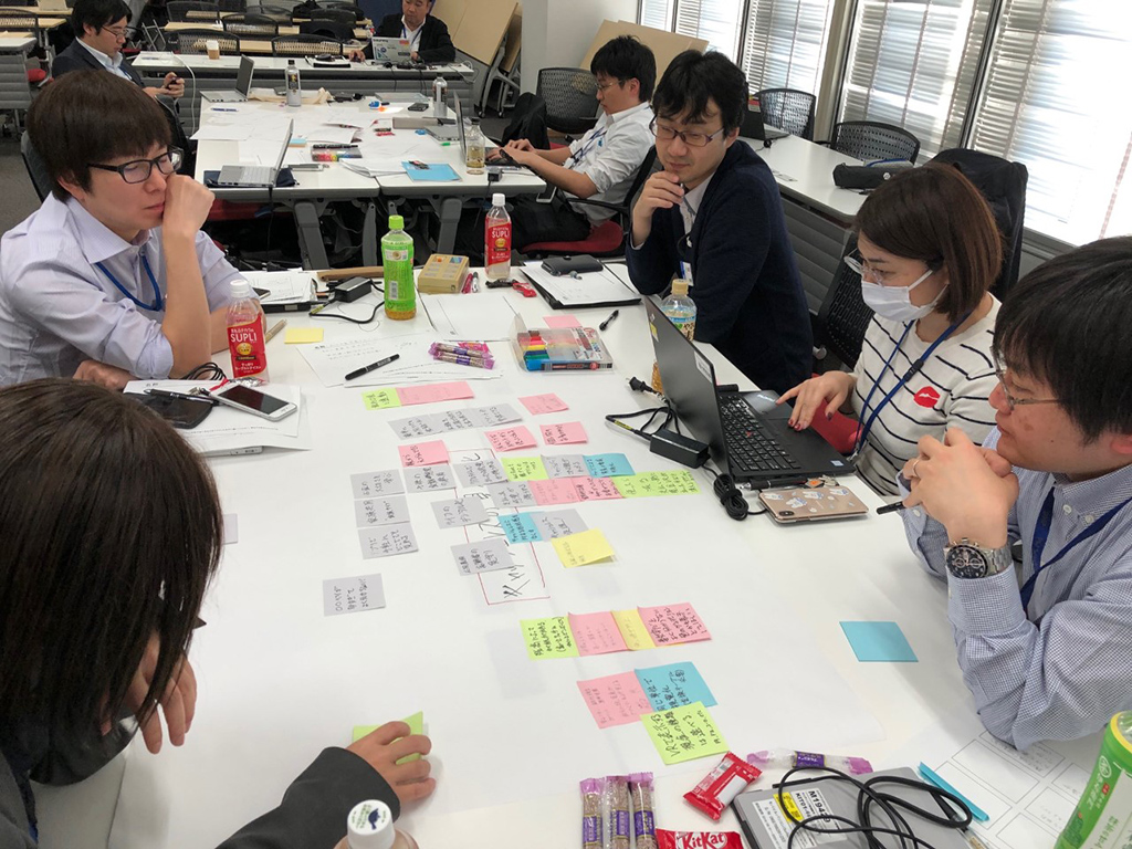 5名の社員が机を囲み、付箋に意見を書きながら話し合う様子