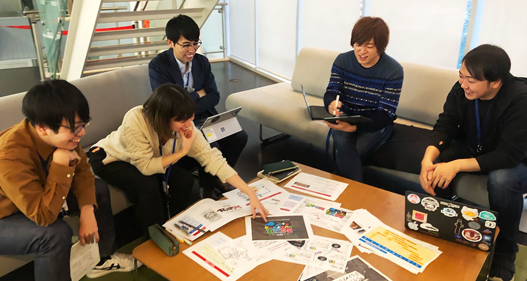 五反田オフィスにて、5名の社員が机上に資料を広げ、話し合う様子